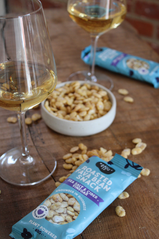 Salt and vinegar roasted fava bean snacks in packets and a bowl with two glasses of bright white wine